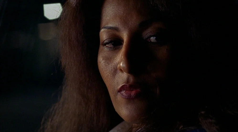 Jackie Brown movie still: closeup — A woman is inside a dark vehicle at night with minimal illumination. She looks past the lens towards an unseen…