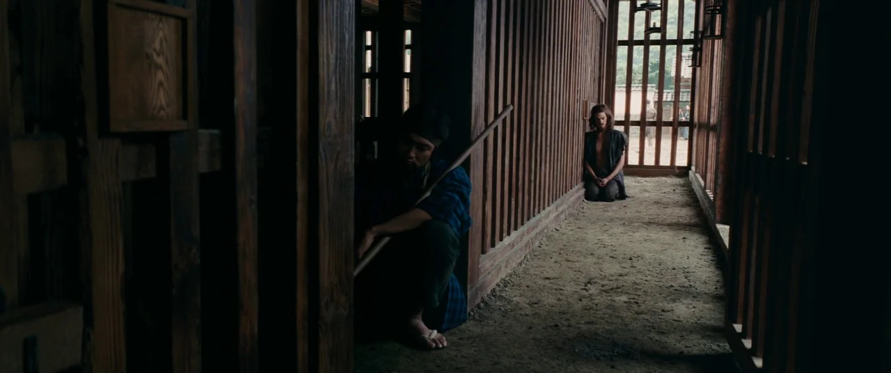 Silence (2016) movie still: wide — A wooden building features slatted vertical walls and a packed earth walkway floor. A man squats in the near…