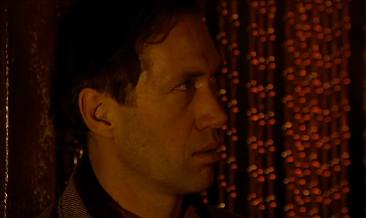 The Serpent’s Egg (1977) movie still: closeup — A dimly lit interior space features a dark wall and an out-of-focus curtain backdrop. A man stands…
