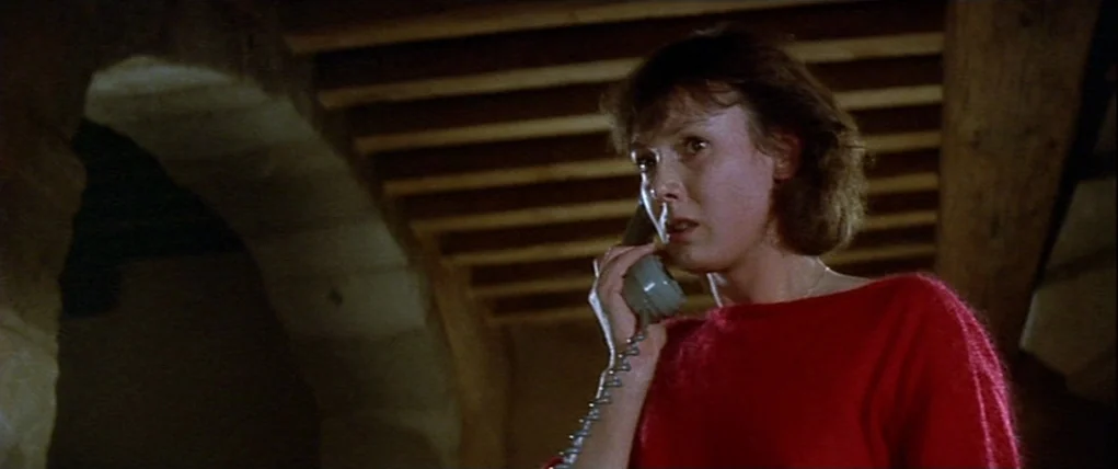 Love Unto Death (1984) movie still: medium — A dimly lit interior space features an arched stone wall and wooden ceiling beams. A woman in a red…