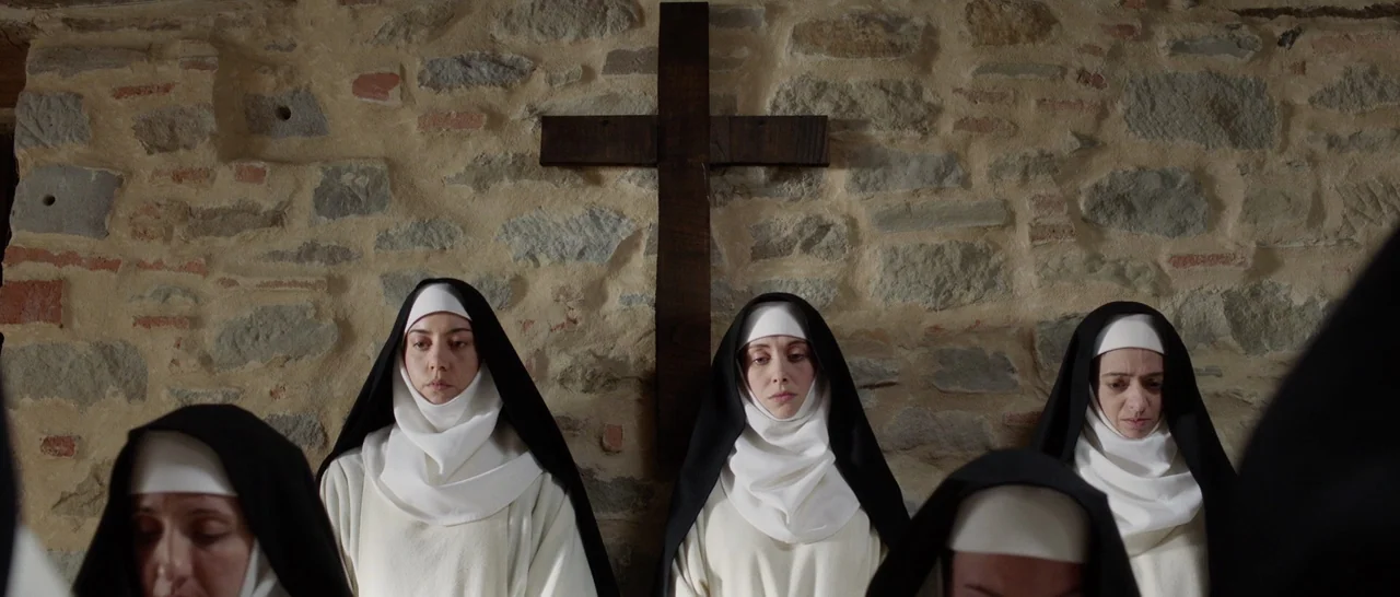 The Little Hours (2017) movie still: medium — A group of women in religious habits stand in front of a stone wall. A dark wooden cross is mounted on…