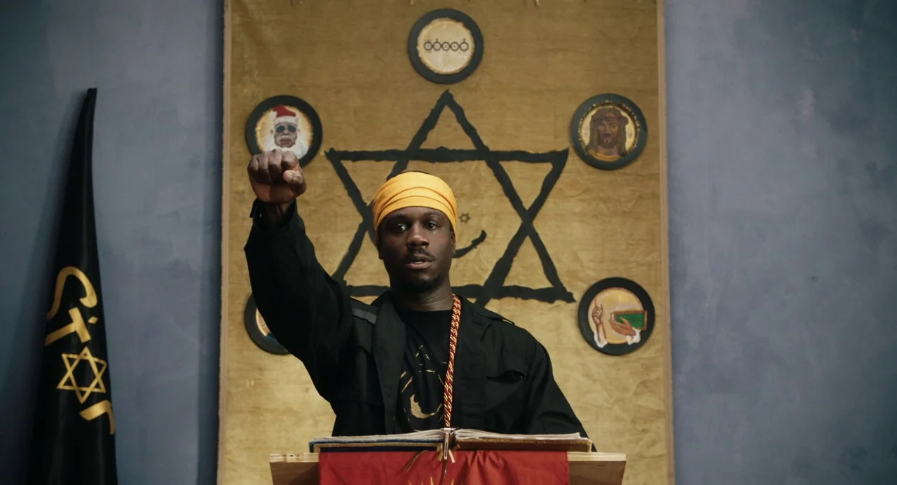 The Day Shall Come (2019) movie still: medium — A man stands behind a wooden lectern in front of a large fabric banner. The banner displays a black…