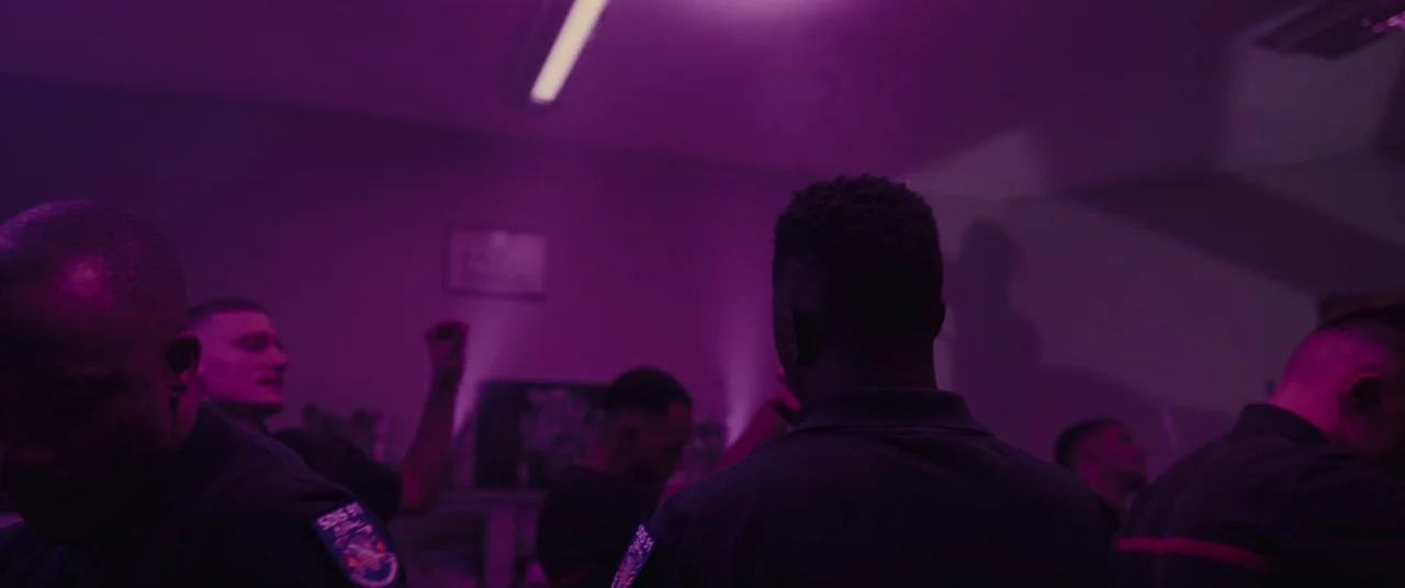 Titane (2021) movie still: medium — An interior space occupied by multiple people wearing dark uniforms under violet light. The men are grouped…