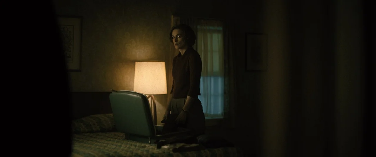 Boston Strangler (2023) movie still: medium — A dimly lit room with patterned wallpaper features a bed and a vintage lamp. A woman packs an open…