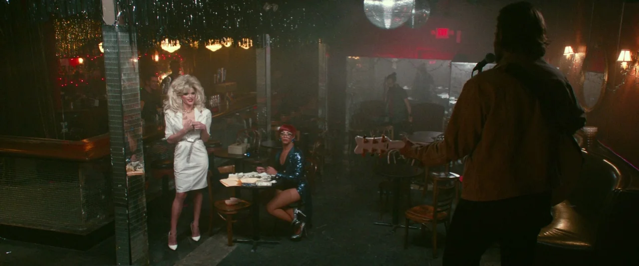 A Star Is Born (2018) movie still: wide — A dimly lit club interior features hazy air, mirrored pillars, and a disco ball. A man with a guitar faces…