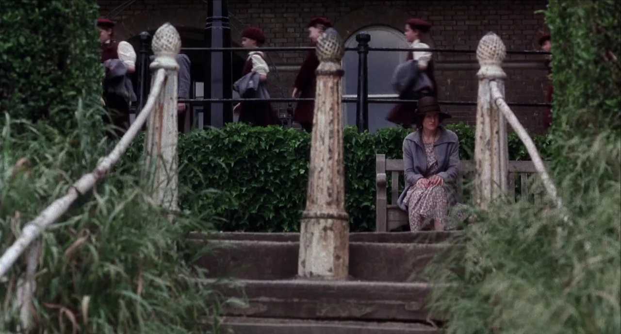 The Hours (2002) movie still: wide — An outdoor stone staircase overlooks a paved area framed by green hedge bushes. A lone woman sits on a wooden…
