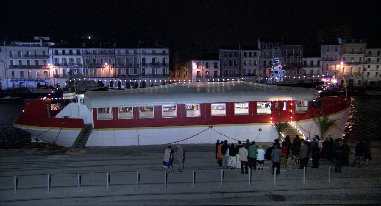 The Secret of the Grain (2007) movie still: wide — A long riverboat painted white and red is docked at a concrete quay at night. A group of people…