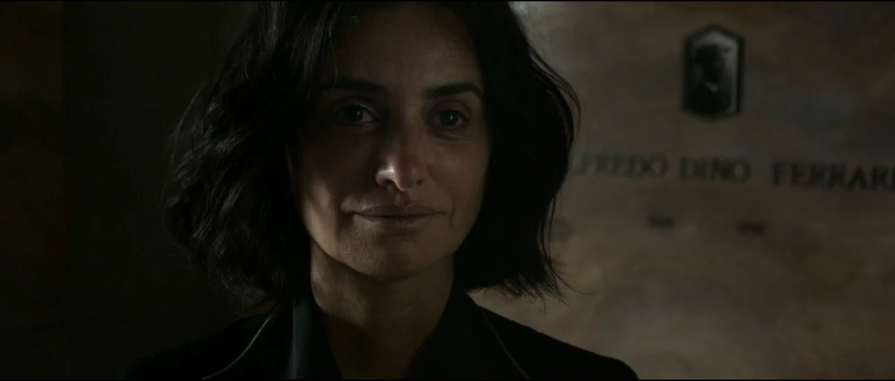 Ferrari (2023) movie still: closeup — A dimly lit indoor mausoleum contains stone wall markers with engraved names and plaques. A woman stands in…