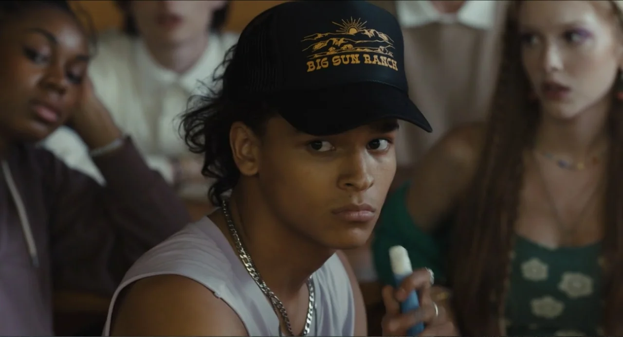 Hell of a Summer (2025) movie still: medium — A group of teenagers sits in a dimly lit indoor educational setting. A young man in the center wears a…