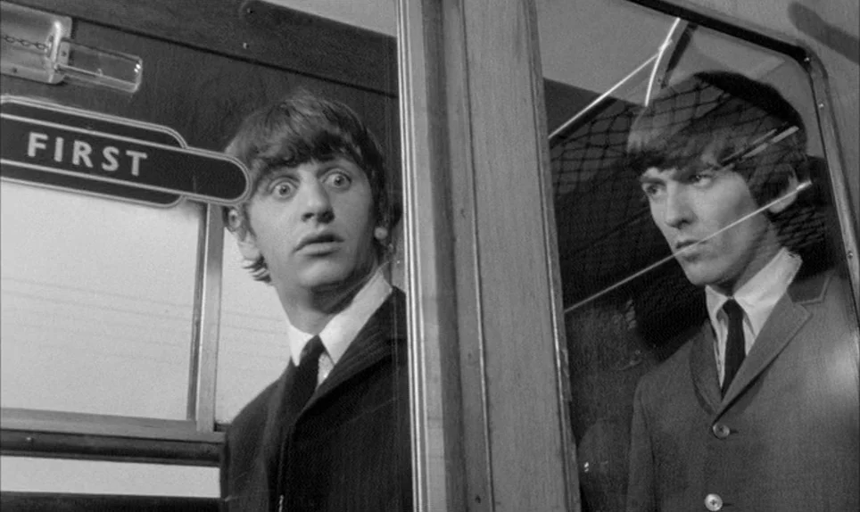 A Hard Days Night (1964) movie still: medium — The interior of a 1960s passenger train carriage with wooden panels and glass partitions. Two young…