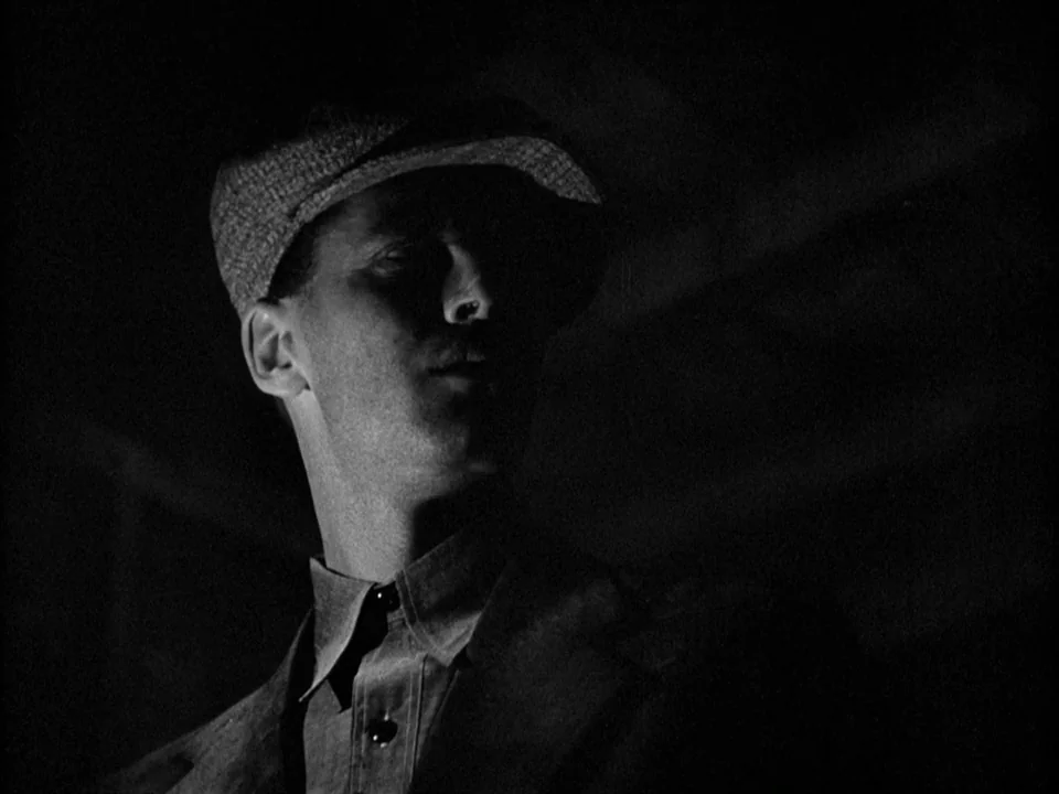 The Grapes of Wrath (1940) movie still: closeup — A man stands in an undifferentiated dark environment with heavy shadows. He wears a textured flat…