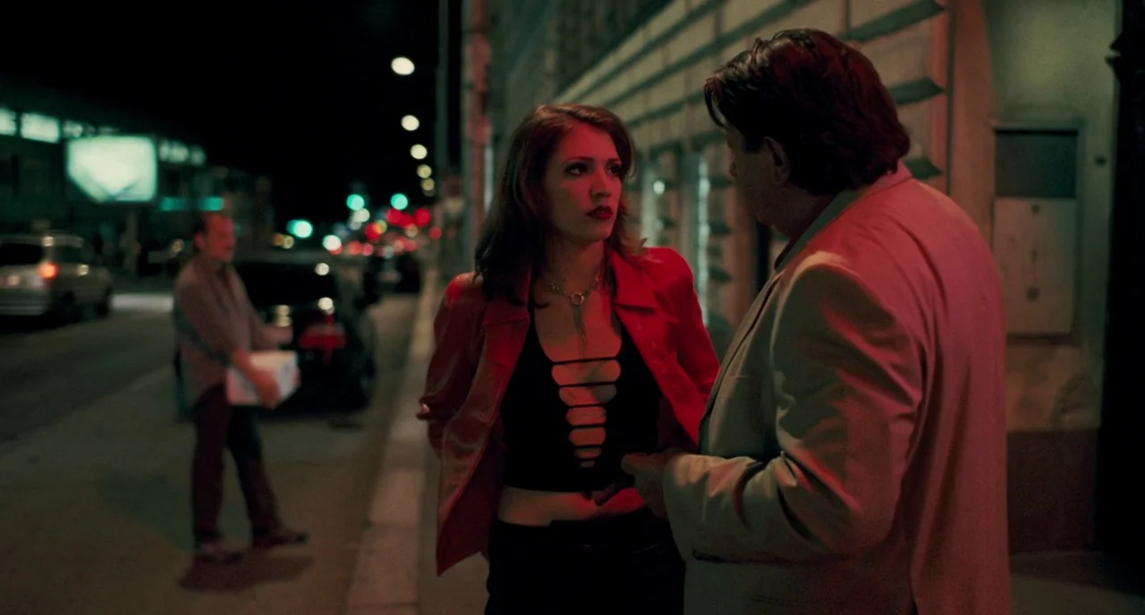 Revanche (2008) movie still: medium — An urban sidewalk at night is illuminated by distant streetlights and glowing neon signs. A woman and a man in…