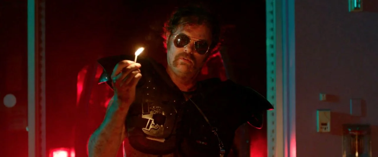 Cooties (2014) movie still: medium — A dark hallway lit by flickering red neon light casts long shadows against the walls. A man wearing black body…