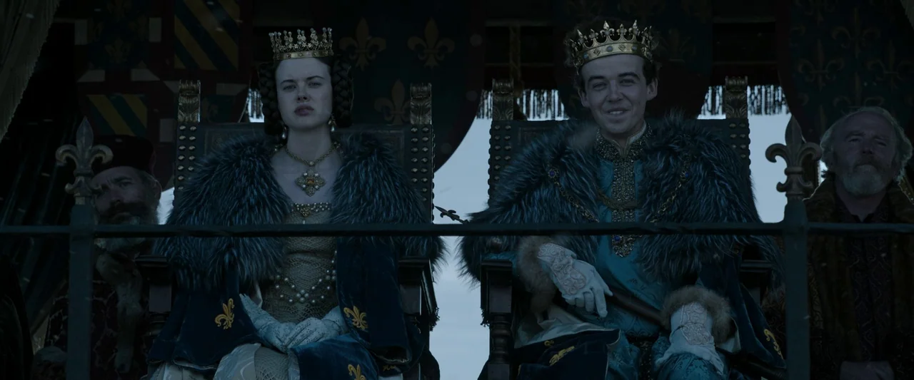 The Last Duel (2021) movie still: medium — A king and queen sit on elaborate wooden thrones placed on an outdoor dais. Dark, fleur-de-lis decorated…