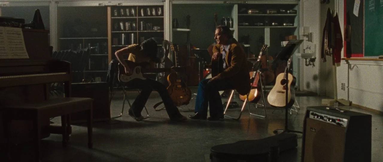 The Runaways (2010) movie still: wide — A music practice room contains shelves filled with instrument cases and various guitars. Two individuals sit…