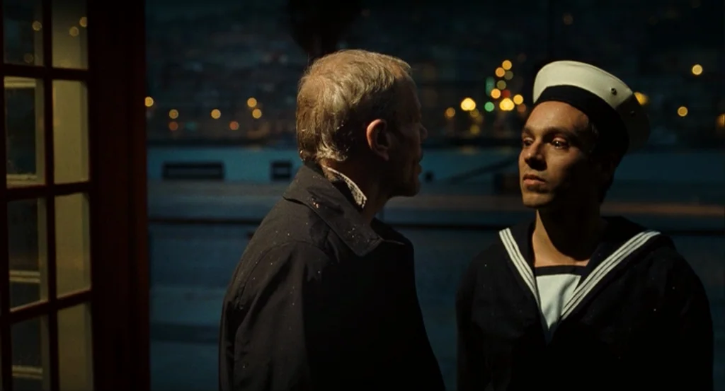 This Night (2008) movie still: medium — A nighttime scene set beside a quiet, dark body of water with city lights. An older man in a dark jacket…