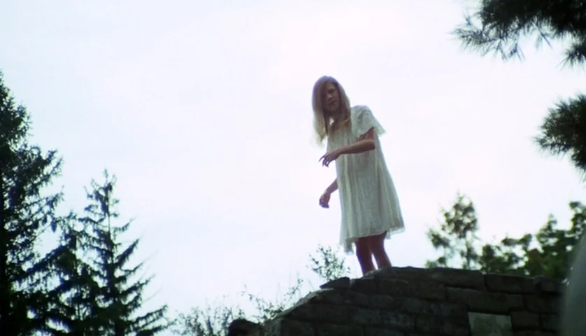 Let’s Scare Jessica To Death (1971) movie still: fullbody — A teenage girl in a white dress stands atop a stone wall outdoors. Pine trees frame the…