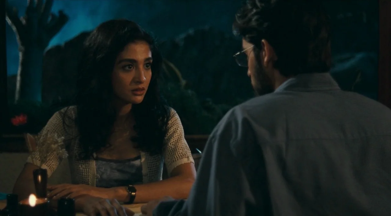 The Other Conquest (1999) movie still: medium — A dimly lit table inside a restaurant at night with blue tones outside. A woman looks at a man who…