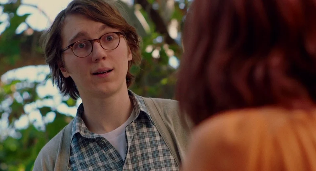 Ruby Sparks (2012) movie still: medium — A man stands outdoors among blurred green trees in a sunny park. He looks toward an unseen person while…