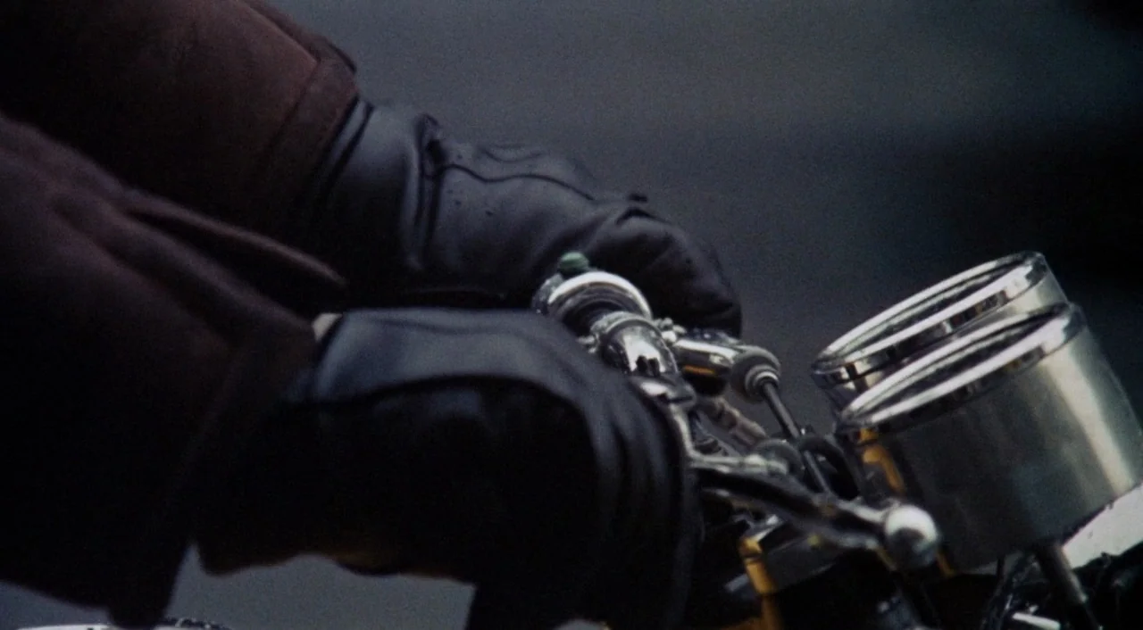 The Iguana with the Tongue of Fire (1971) movie still: closeup — A close view of black leather gloves resting on motorcycle handlebars with metallic…