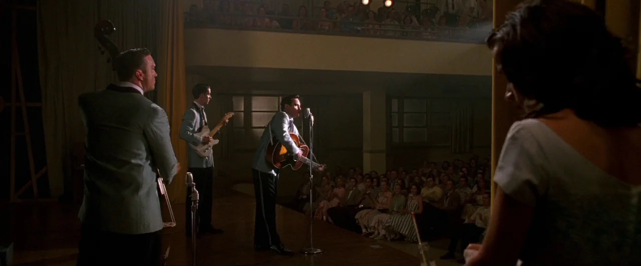 Walk The Line (2005) movie still: wide — A 1950s auditorium features a raised wooden performance space with a large seated audience. Three men play…