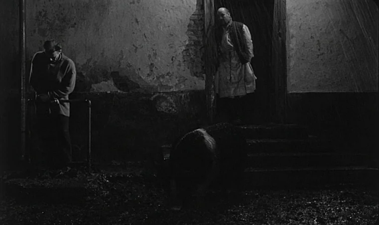 Sátántangó (1994) movie still: wide — A dark, gritty alleyway features a weathered brick wall and a stone staircase. One man leans against a railing…