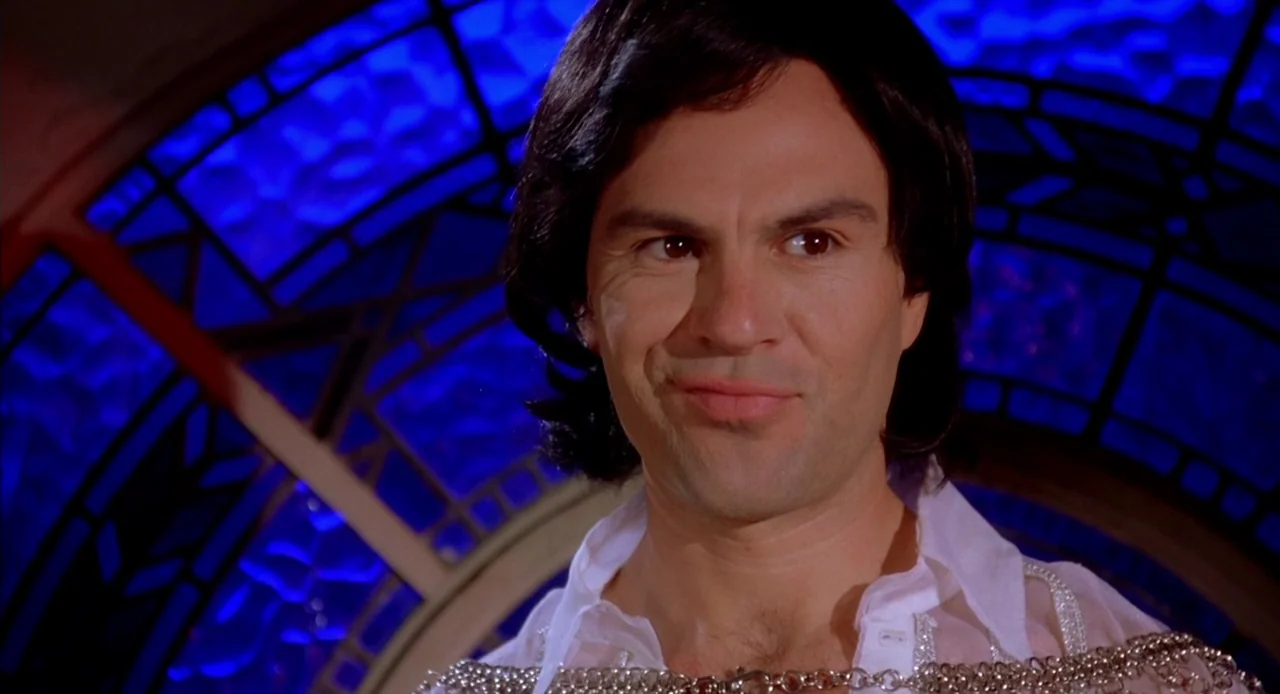 Viva (2007) movie still: closeup — A dimly lit space features an illuminated arched blue stained-glass wall. A man with dark shoulder-length hair…