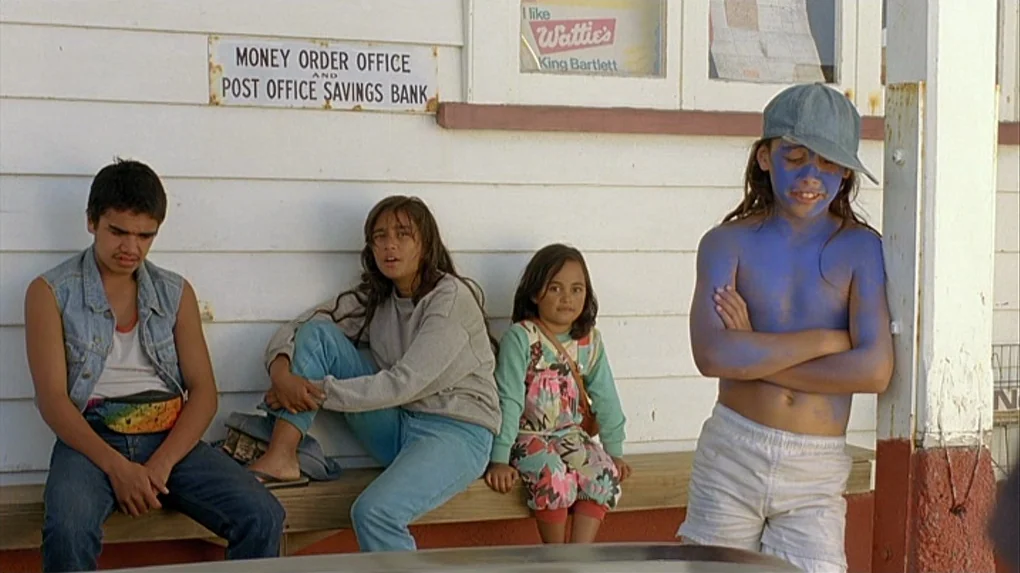 Boy (2010) movie still: fullbody — A group of children sits on a wooden bench against a white painted wall. The building wall features a metal sign…