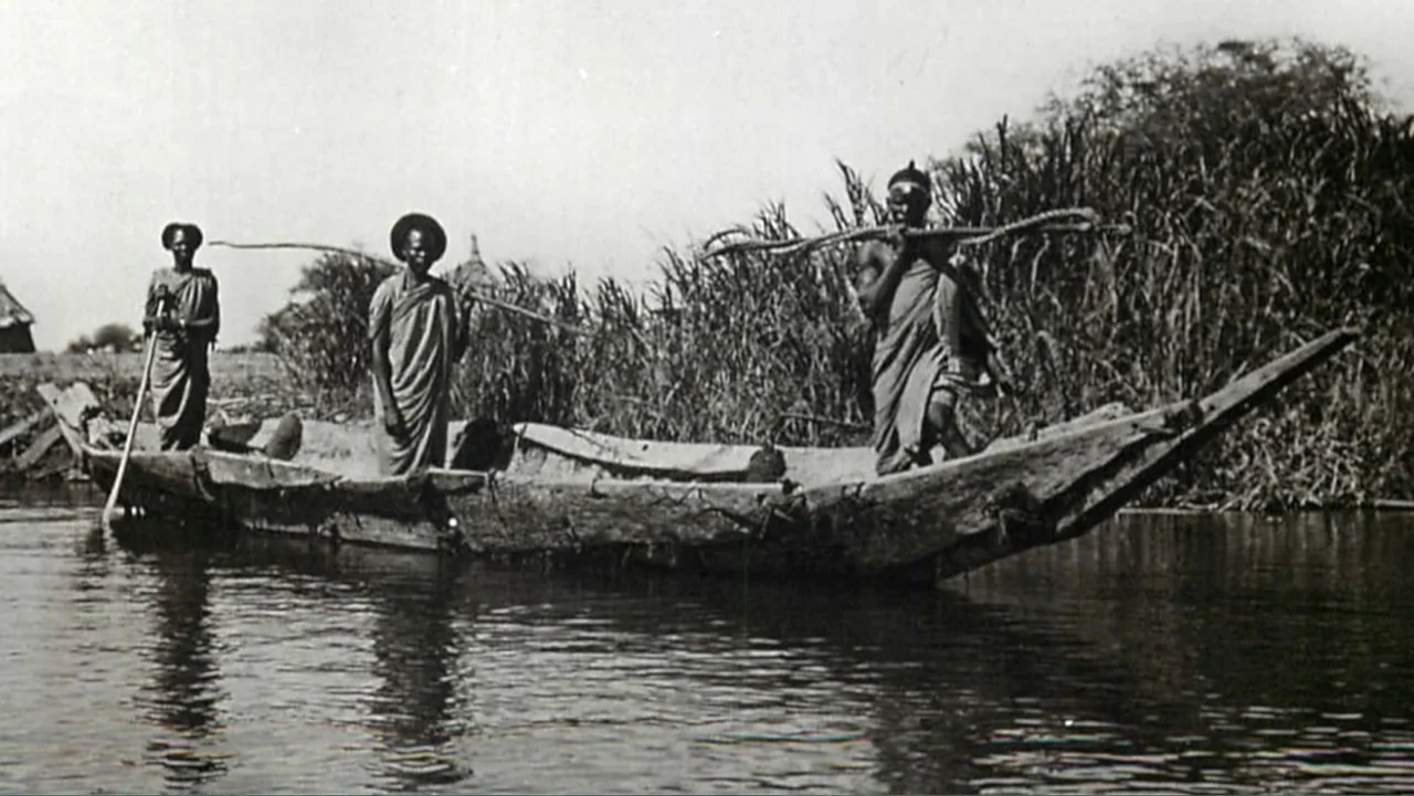 The Atlantis Puzzle (2024) movie still: wide — A wooden dugout canoe travels along a calm waterway bordered by dense tall reeds. Three men stand in…