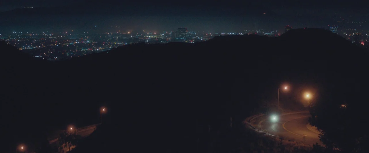 Gemini (2017) movie still: wide — A vast night view shows a winding mountain road illuminated by scattered yellow streetlamps. In the distance, a…