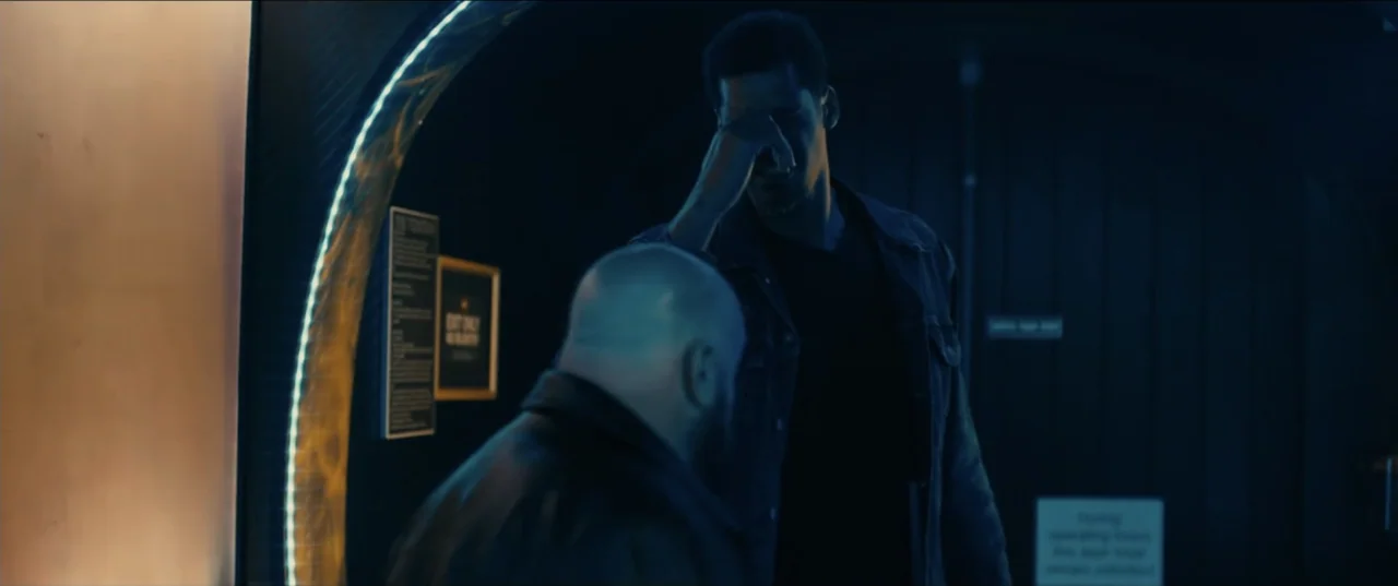 Guns Up (2025) movie still: medium — A dimly lit doorway at a night venue features two men interacting near a neon sign. The taller man touches his…