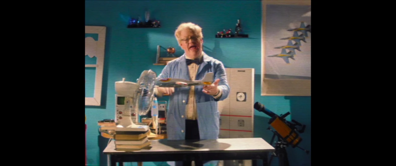 Linoleum (2022) movie still: medium — A scientist stands before a desk in a blue room with scientific instruments. He holds a model airplane while…