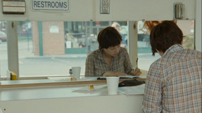 Wendy and Lucy (2008) movie still: medium — A roadside diner booth features a mirror reflecting two people sitting across from each other. One woman…
