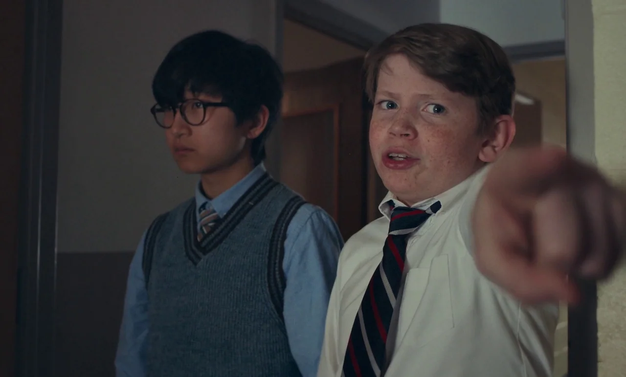 The Holdovers (2023) movie still: medium — A dimly lit indoor hallway with plain walls and an open doorway behind subjects. Two boys stand together…