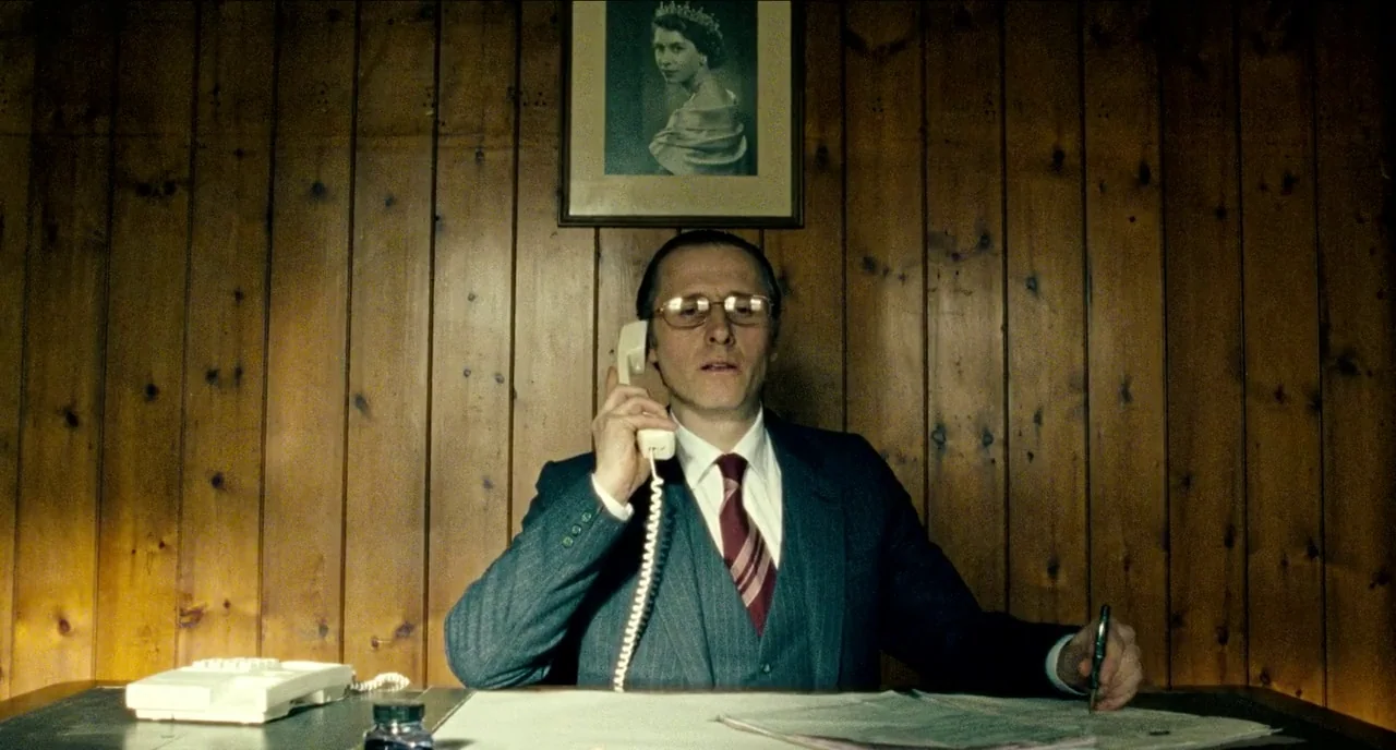 Bronson (2008) movie still: medium — A man sits at a desk in front of wall-mounted vertical wood paneling. Queen Elizabeth II is depicted in a…