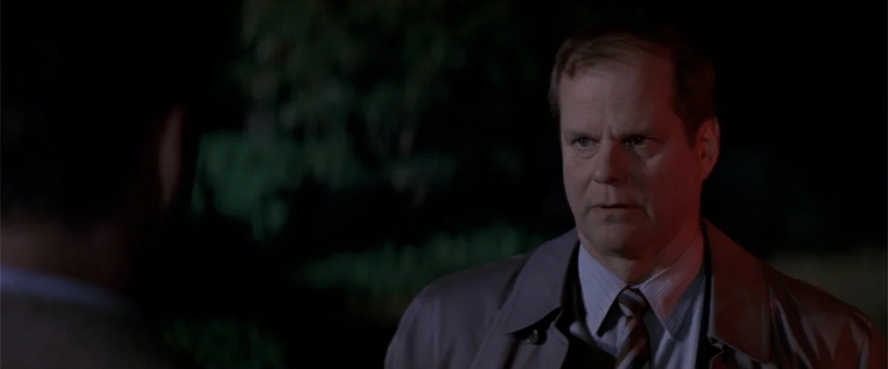Angst (2017) movie still: medium — A dark outdoor space with dense foliage visible in the out-of-focus background. A middle-aged man in a trench…