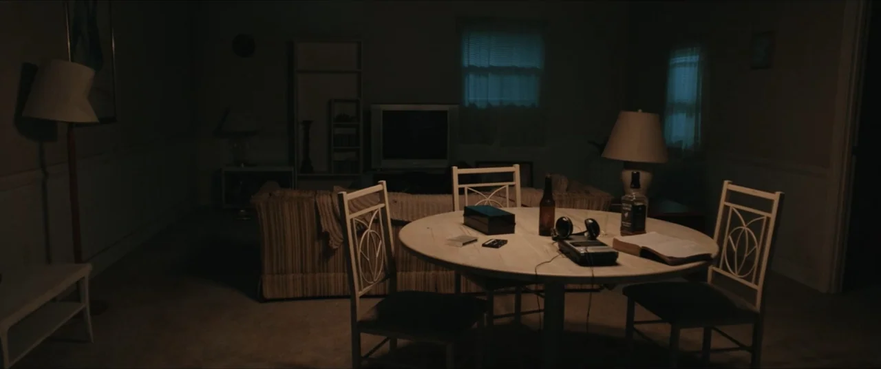 Possessions (2024) movie still: wide — A dimly lit 1990s dining room features a round table with scattered personal items. The surrounding space…