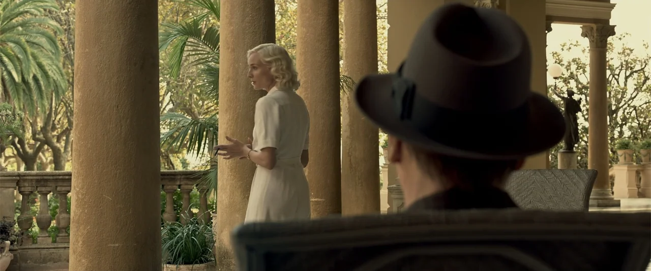 Marlowe (2023) movie still: medium — A stone-columned veranda overlooks a lush green landscape with ornamental statues and palm trees. A woman in a…