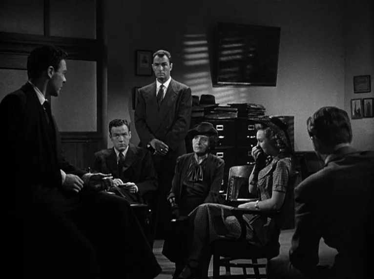 White Heat (1949) movie still: wide — A dark office setting features a group of people sitting around a central figure. The room contains filing…
