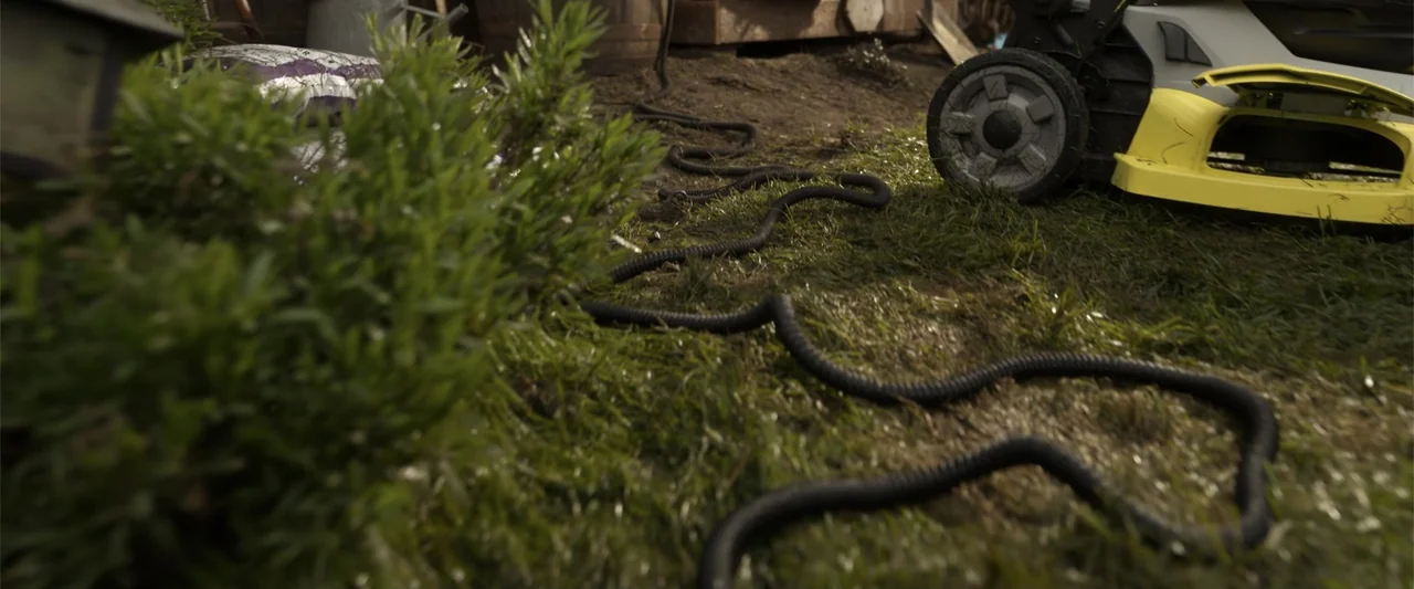 Final Destination Bloodlines (2025) movie still: medium — A neglected backyard patch shows grass and a black coiled hose near a lawnmower. The…