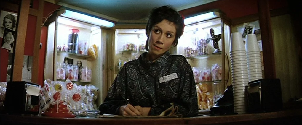 Anguish (1987) movie still: medium — A small retail shop counter is surrounded by shelves stocked with jars of candy. A woman sits behind the…