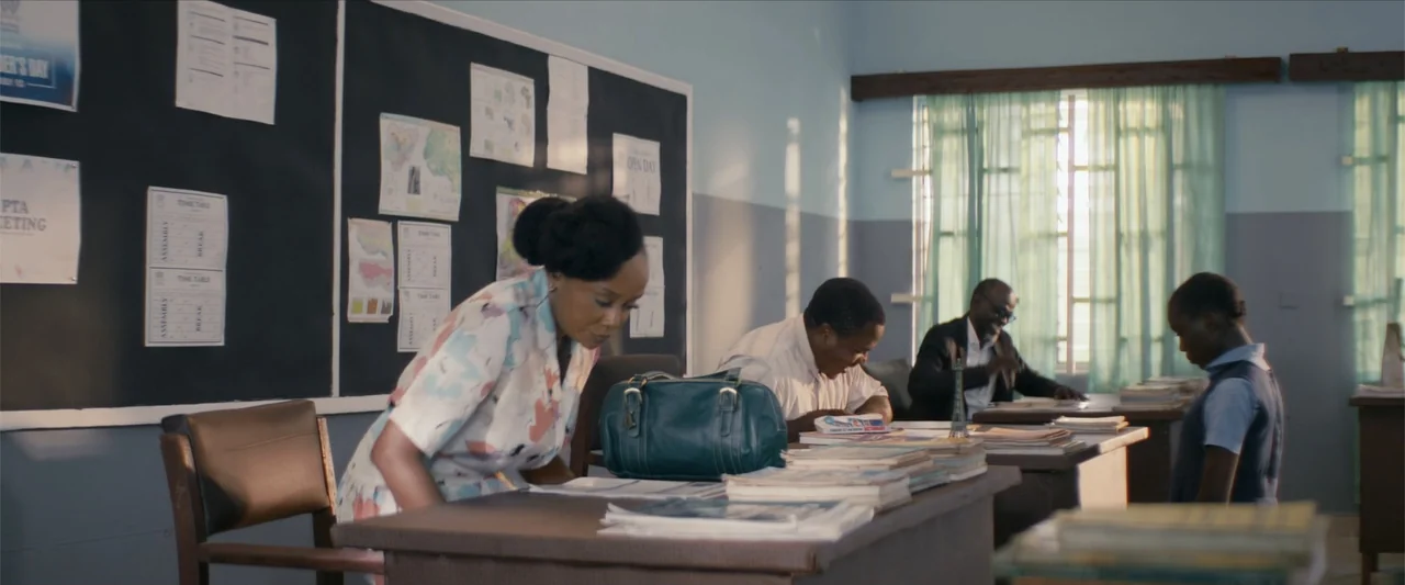 Ms. Kanyin (2025) movie still: medium — A school staff room features desks, a black chalkboard with notices, and large windows. A woman wearing a…