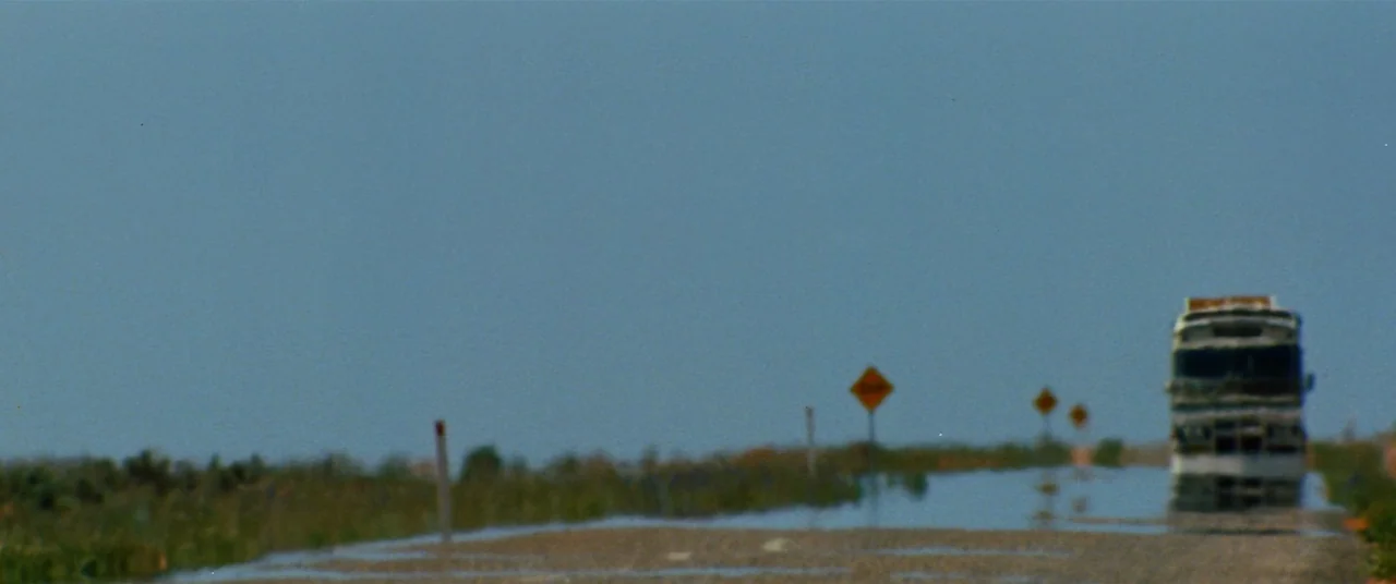 Razorback (1984) movie still: wide — A flat, arid landscape stretches toward a clear blue sky above a paved highway. A large transport truck…