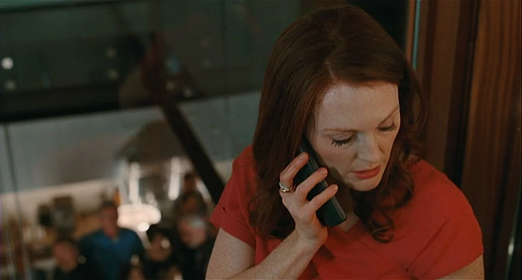 Chloe (2009) movie still: closeup — A woman stands near a glass staircase in a modern home interior. She is focused on a conversation while holding…