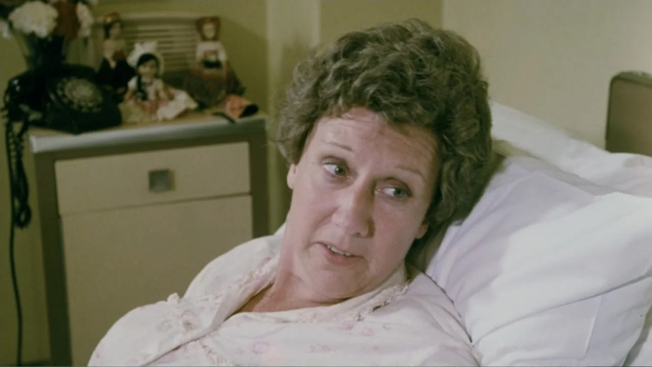 Aunt Mary (1979) movie still: medium — A sterile hospital room features a bed against a plain wall and a side table. An older woman in a medical…