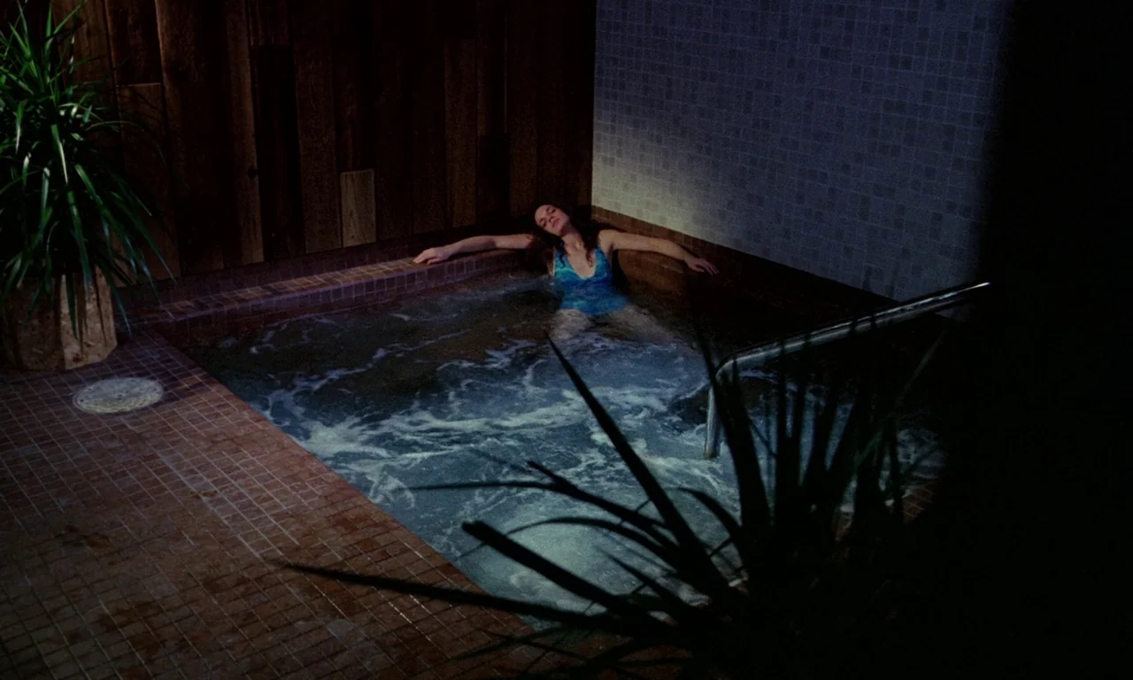Rabid (1977) movie still: wide — A tiled indoor hot tub area is enclosed by wood-paneled walls and a potted plant. A woman rests with her head back…