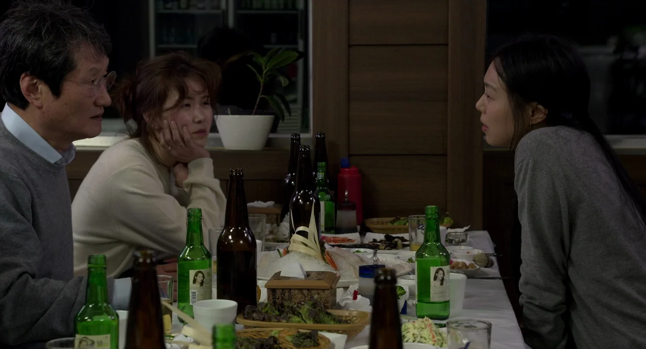 On The Beach At Night Alone (2017) movie still: medium — A restaurant dining table filled with plates of food and numerous green soju bottles. Two…