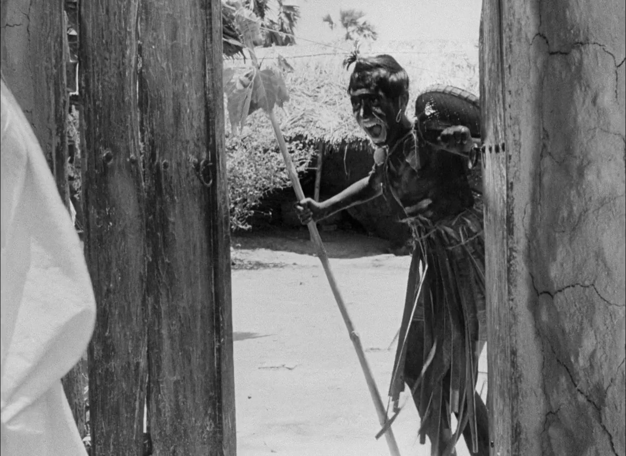 Aparajito (1956) movie still: fullbody — A rustic doorway frames a boy standing in a clearing with thatched roof structures. The boy holds a long…