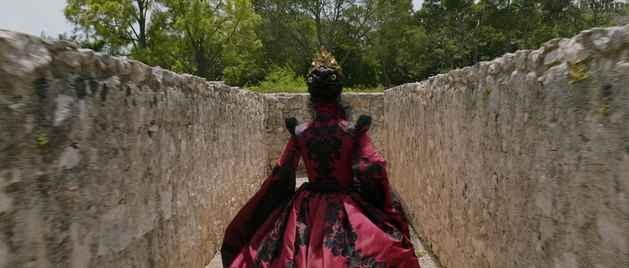 Tale of Tales (2015) movie still: fullbody — A narrow passage flanked by rough-hewn stone walls leads to a tree-lined area. A woman in an elaborate…