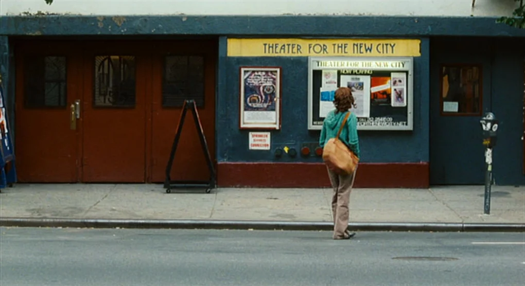 The Savages (2007) movie still: fullbody — The exterior of a community performing arts venue shows red double doors and posters. A person wearing a…